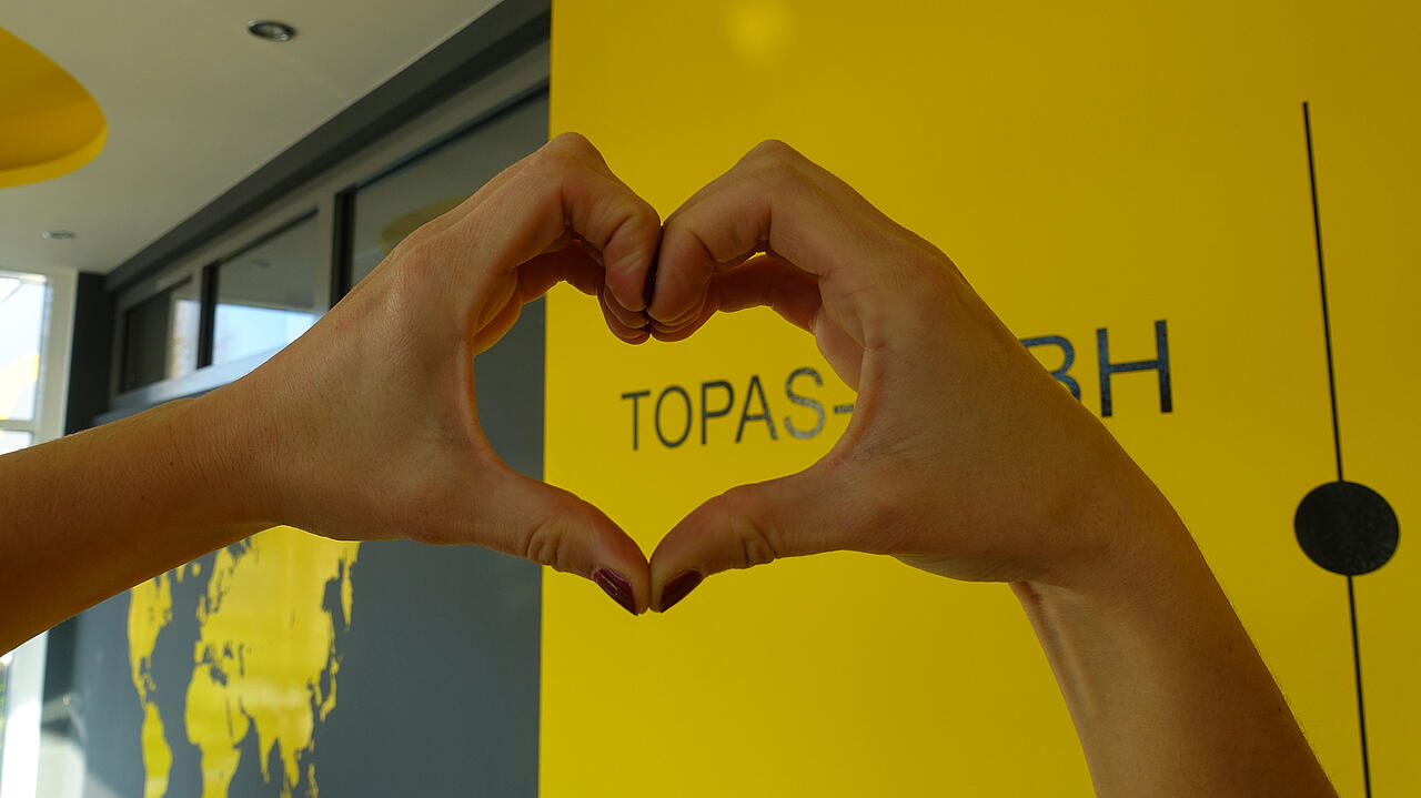 Zona de entrada de Topas GmbH con un mapamundi y el nombre de la empresa; delante, unas manos que forman un corazón frente al nombre «Topas».