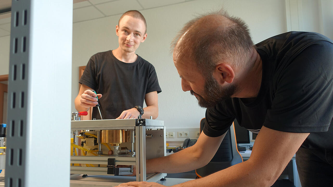 El aprendiz levanta la vista de la pieza de trabajo; en primer plano, otro ingeniero está trabajando