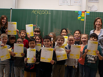 Una clase de colegio, con los alumnos sonriendo, reunida delante de la pizarra con sus certificados en la mano y la palabra «Aerosol» escrita en la pizarra.