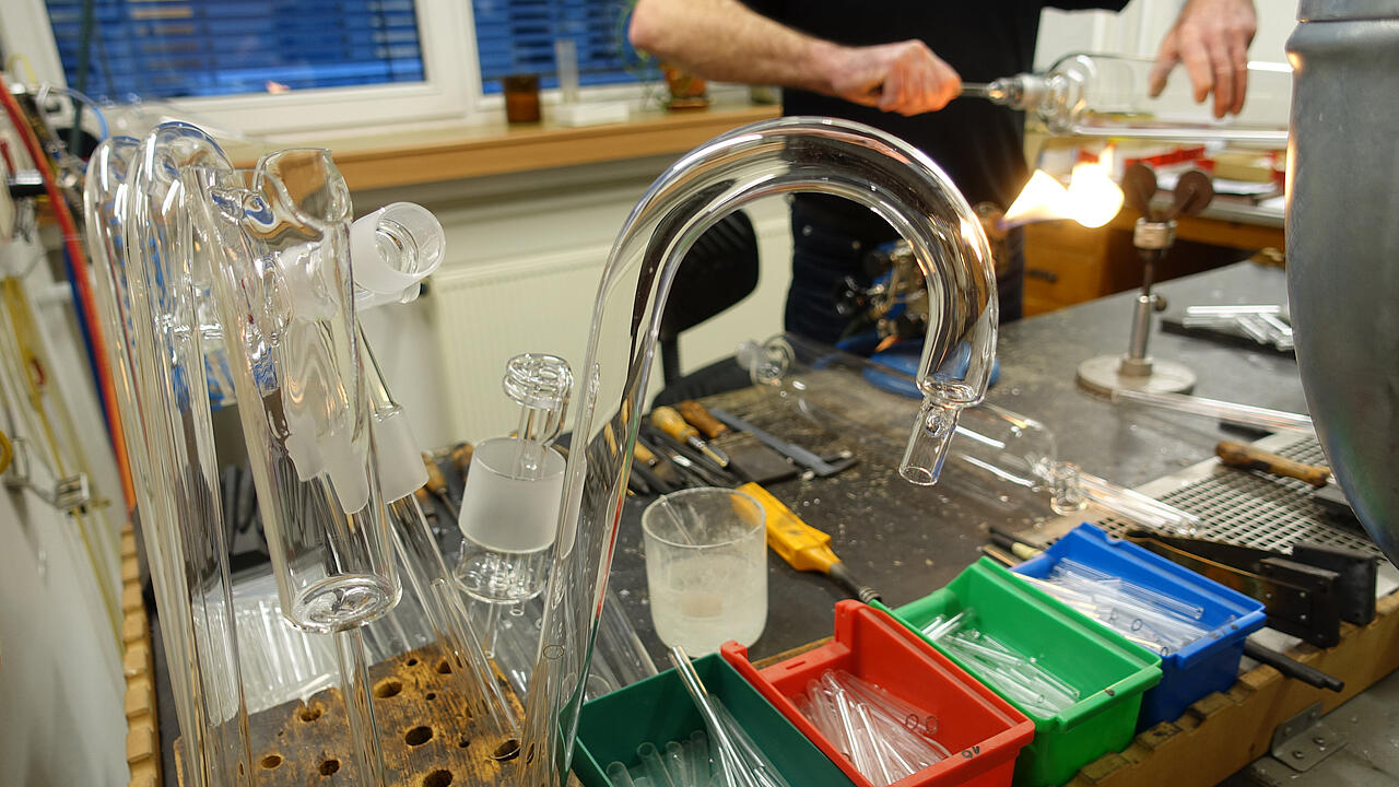 [Traduire en anglais :] Souffleur de verre au travail, avec au premier plan des objets en verre et des outils